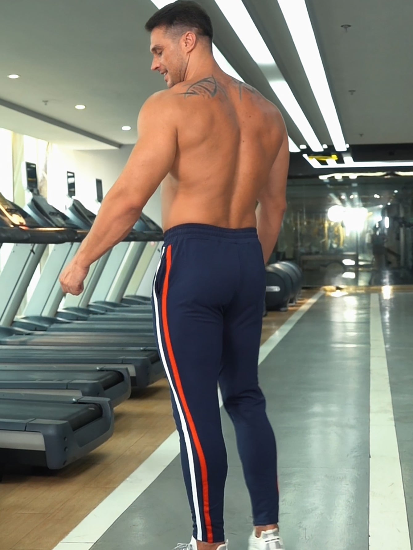 Back view of man wearing Anvtt navy blue side-stripe tapered jogger 
pants in gym – men's slim fit training bottoms with elastic waistband, 
perfect for leg day and gym sessions